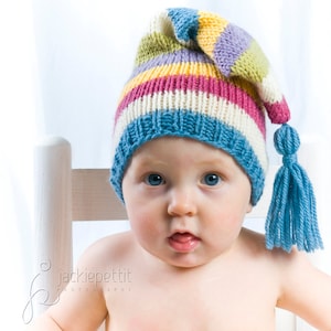 May include: A baby wearing a colorful striped knitted hat with a blue tassel. The hat has stripes of pink, yellow, blue, and green.