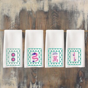 May include: Four white kitchen towels with embroidered green squares containing Chinese symbols and images. The symbols include a dragon, two circles, a character, and two vertical lines.