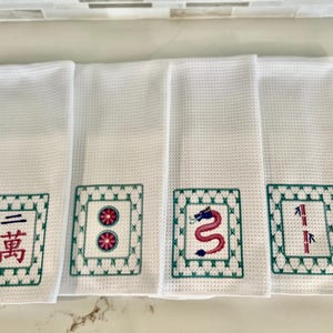 May include: Four white kitchen towels with embroidered green and red Chinese mahjong tiles. The tiles feature a dragon, bamboo, circle, and a character.