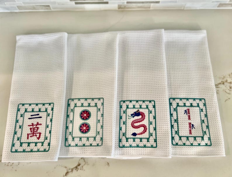 May include: Four white kitchen towels with embroidered green and red Chinese mahjong tiles. The tiles feature a dragon, bamboo, circle, and a character.