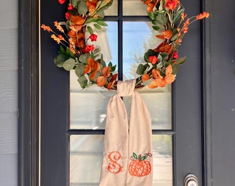May include: A circular autumn wreath featuring orange and red foliage, green leaves, and thin branches. A beige fabric ribbon hangs from the bottom, embroidered with an orange letter S and an orange pumpkin graphic with green leaves on the stem.