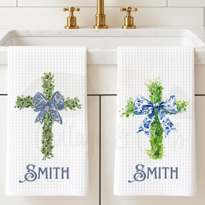 May include: Two white waffle-weave hand towels with a cross design. The crosses are made of green foliage and adorned with blue bows. The word "SMITH" is printed in blue script below each cross. Gold faucet fixtures are visible in the background.