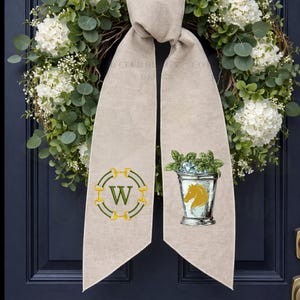 May include: A beige door sash with a green and gold letter "W" design and a bucket with a horse head illustration. The sash is hanging in front of a wreath of white flowers and green leaves.