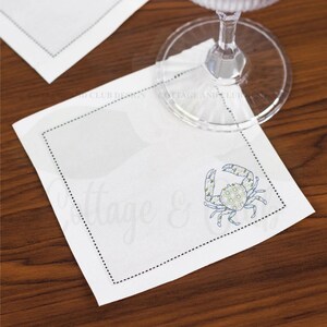 May include: White square linen cocktail napkin with a black dotted border and an embroidered crab design in blue, green, and yellow. A clear wine glass sits above the napkin. The napkin is on a wooden surface.