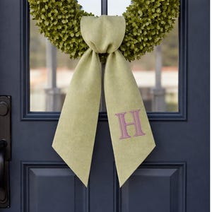 May include: A green wreath with a light green bow and the letter "H" embroidered in pink. The wreath is hanging on a navy blue door. The bow is made of a textured fabric.