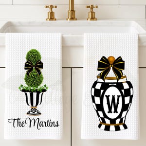 May include: Two white waffle-weave hand towels with decorative designs. One features a topiary with a black bow, the other a black and white checkered vase with a black bow and the letter "W". The text "The Martins" is on one towel.