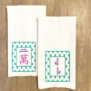 May include: Two white kitchen towels with embroidered Chinese characters and bamboo designs. The first towel has a green square frame with the Chinese character for "three" and the word "bamboo" in a smaller square inside. The second towel has a green square frame with a stylized image of two bamboo stalks with leaves.