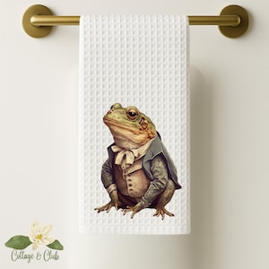 May include: A white waffle weave kitchen towel with a green and brown frog wearing a blue and white suit and a white bow tie.