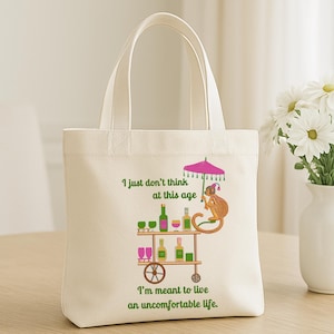 May include: A white tote bag with a green and pink design featuring a monkey sitting at a bar cart with drinks and an umbrella. The text on the bag reads "I just don't think at this age I'm meant to live an uncomfortable life."
