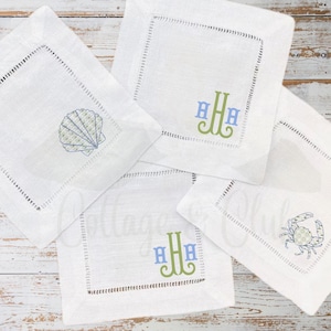 May include: Four white square coasters with embroidered designs. One features a seashell, another a crab, and two display a monogram in blue and green. Each coaster has a stitched border.