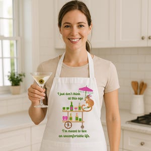 May include: White apron with a monkey holding an umbrella and a bar cart with drinks. The text on the apron reads "I just don't think at this age I'm meant to live an uncomfortable life."
