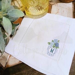 May include: White linen napkin featuring an embroidered vase with blue and green hydrangeas. The vase has a blue and white pattern and the letter "M". The napkin has a stitched border.