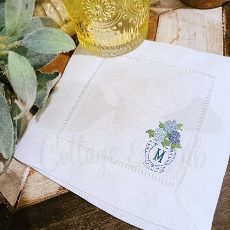 May include: White linen napkin featuring an embroidered vase with blue and green hydrangeas. The vase has a blue and white pattern and the letter "M". The napkin has a stitched border.