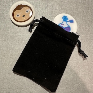 May include: Two small ceramic tokens in a black velvet drawstring bag. One token features a cartoon monkey face, the other a cartoon potion bottle.