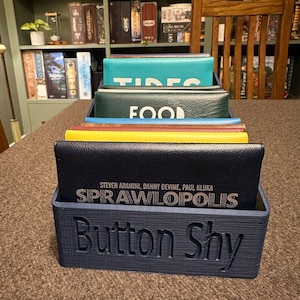 Könnte beinhalten: Eine dunkelblaue, 3D-gedruckte Organizer-Box mit dem Text "Button Shy" auf der Vorderseite, die mehrere Brettspiele enthält. Die Spiele haben verschiedenfarbige Cover, darunter Türkis, Grün, Gelb und Marineblau. Das Spiel "Sprawlopolis" ist sichtbar.