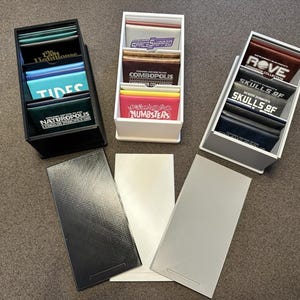 May include: Three white, black, and gray card holders filled with colorful cards. The cards have text such as "TIDES", "NUMBSTERS", and "SKULLS OF". Three matching colored card holder covers are also present.