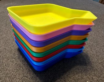 May include: A stack of eight colourful plastic trays, each with a slightly different shape. The trays are arranged in a rainbow order, starting with yellow at the top and ending with blue at the bottom.