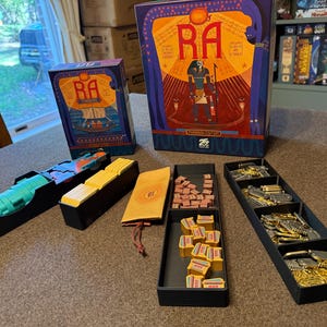 May include: The board game "Ra" is displayed, featuring a large box with the game's title and an illustration of an Egyptian pharaoh. Various game components, including tokens and card holders, are arranged around the box. The game's theme is ancient Egypt.