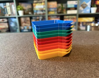 May include: A stack of colourful, plastic game trays in blue, green, red, orange, and yellow. The trays are stacked on top of each other, with the colours arranged in a rainbow order. The trays are likely for organising game pieces.