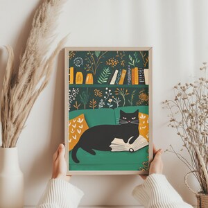 May include: A framed art print featuring a black cat lounging on a green sofa, reading a book. The background shows a bookshelf with books and decorative vases. The print has a botanical pattern in shades of green, orange, and white.