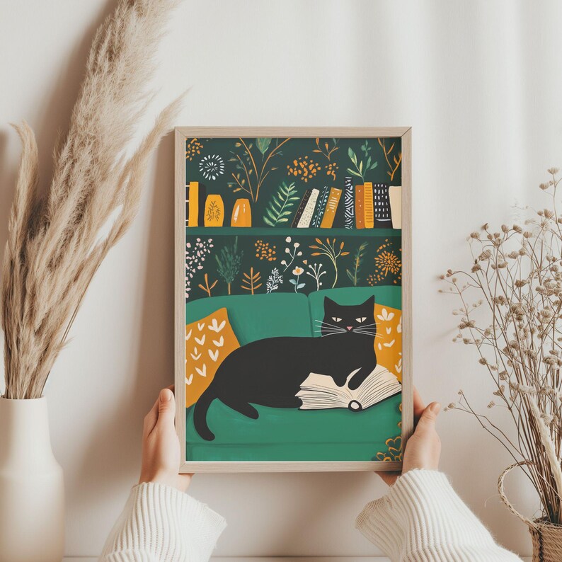 May include: A framed art print featuring a black cat lounging on a green sofa, reading a book. The background shows a bookshelf with books and decorative vases. The print has a botanical pattern in shades of green, orange, and white.