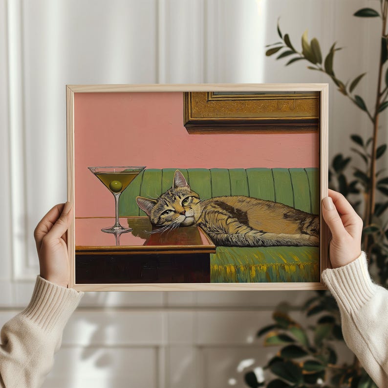 A framed painting of a tabby cat lying on a green couch with a martini glass on a table in front of it. The background is a light pink wall with a gold picture frame on the wall.