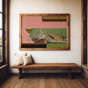 A framed painting of a tabby  cat lying on a green couch with a martini glass on a table in front of it. The background is a light pink color.