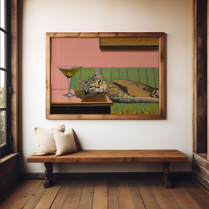 A framed painting of a tabby  cat lying on a green couch with a martini glass on a table in front of it. The background is a light pink color.