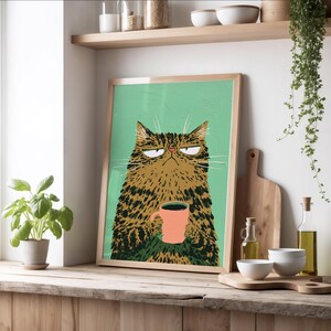 May include: A framed wall art print featuring a stylized illustration of a grumpy brown tabby cat with long whiskers holding a solid pink mug against a solid light green background. The cat has narrow eyes and a stern expression.