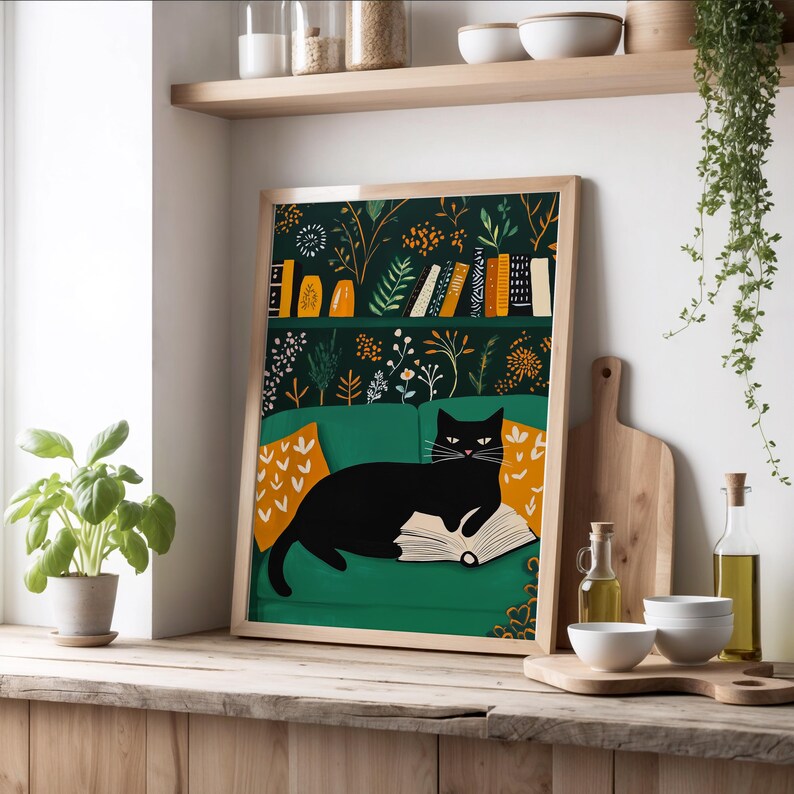 May include: A framed art print featuring a black cat lounging on a green couch, reading a book. The background has a dark green floral pattern with a bookshelf. The couch has orange pillows with white arrow designs.