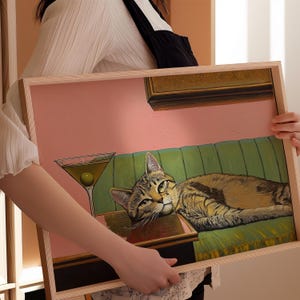 A framed painting of a cat lying on a green couch with a martini glass on a table in front of it. The background is a pink wall with a gold picture frame on it.