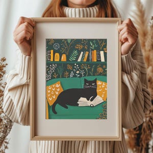 May include: A framed art print featuring a black cat lounging on a green sofa with a book. The background shows a bookshelf and floral wallpaper. Yellow pillows add color to the scene. The frame is a light wood color.
