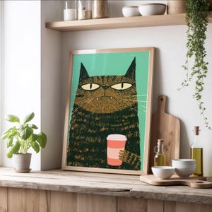 May include: A framed rectangular wall art print featuring a stylized illustration of a brown tabby cat with a grumpy expression, holding a pink coffee cup against a solid mint green background. The cat has pointed ears and visible whiskers.
