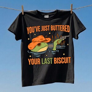 Può includere: Una t-shirt nera a maniche corte con la grafica di una rana verde che indossa un cappello da cowboy arancione e impugna un revolver. Il testo recita YOU'VE JUST BUTTERED YOUR LAST BISCUIT in lettere arancioni e verdi, circondato da stelle.