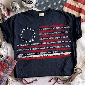 May include: A dark navy blue t-shirt featuring a graphic of a circle of thirteen white stars and the text: WE HOLD THESE TRUTHS TO BE SELF EVIDENT, THAT ALL MEN ARE CREATED EQUAL. THAT THEY ARE ENDOWED BY THEIR CREATOR WITH CERTAIN UNALIENABLE RIGHTS.