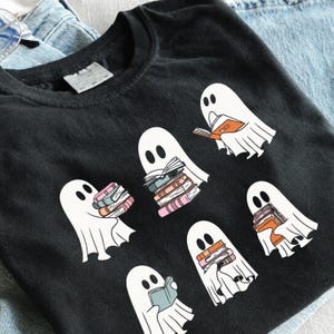 May include: A black t-shirt featuring a graphic print of five white ghosts. Each ghost is depicted interacting with stacks of colorful books or reading an open book. The ghosts have simple black oval eyes and are arranged across the front of the shirt.