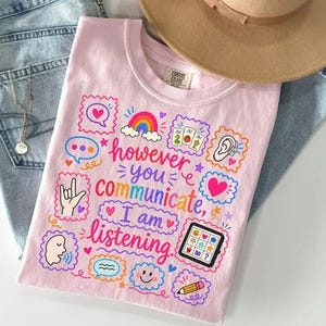 Könnte beinhalten: Ein hellrosa T-Shirt mit dem Text "however you communicate, I am listening" in rosa Schreibschrift. Das Design enthält bunte Illustrationen eines Regenbogens, Sprechblasen, eines Hörgeräts, einer Gebärdensprache-Hand und eines Tablets.