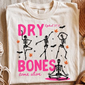 May include: A cream-colored t-shirt featuring a graphic print of four black skeletons in various poses. The text reads "DRY BONES" in large pink letters, "come alive" in smaller pink script, and "Ezekial 37:5" in pink. Orange maple leaves and black bats are scattered throughout.