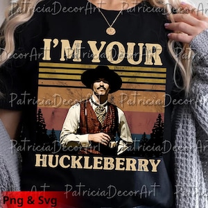 Puede incluir: Camiseta negra con un gráfico vintage de un hombre con sombrero de vaquero y chaleco, con el texto "I'M YOUR HUCKLEBERRY". El diseño incluye un fondo de puesta de sol y árboles.