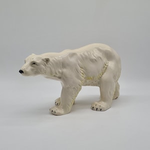 May include: A ceramic polar bear ornament in a walking pose. The bear is mainly off-white with black detailing on the nose and paws. The figurine has textured fur and is set against a plain white background.