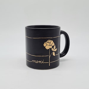 Könnte beinhalten: Schwarze Keramik-Kaffeetasse mit einem goldenen Rosenmotiv und dem Wort "merci" auf der Vorderseite.