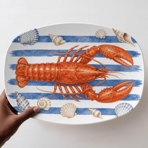 May include: An oval serving platter featuring a central illustration of a bright orange lobster. The background consists of horizontal blue brushstroke stripes interspersed with various detailed illustrations of seashells in shades of white and tan.
