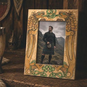 May include: A decorative wooden picture frame with a detailed, ornate design featuring two dragon-like creatures. The frame surrounds a photo of a person in a kilt. A drinking horn and shield are in the background.