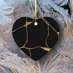 May include: Black heart-shaped ornament with gold-colored crack-like patterns. The ornament hangs from a gold-colored string. The background features a frosty, icy pattern.