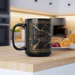 May include: Black ceramic mug with a large handle. The mug features the text "Beautiful Broken" in gold lettering on a black marble-like background. The mug is sitting on a wooden surface.
