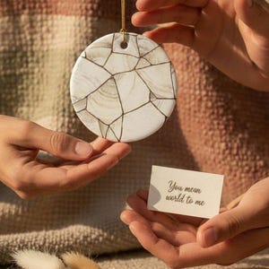 May include: A circular white ceramic ornament featuring a gold-lined mosaic pattern suspended by a thin gold cord, accompanied by a small rectangular card with the printed text "You mean world to me" and a cluster of dried beige grass stems.
