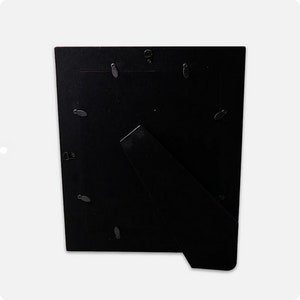 May include: A black picture frame back with a built-in easel stand. The back is made of a dark material and features several small, silver-colored clips to hold a picture in place. The easel is angled to allow the frame to stand upright.