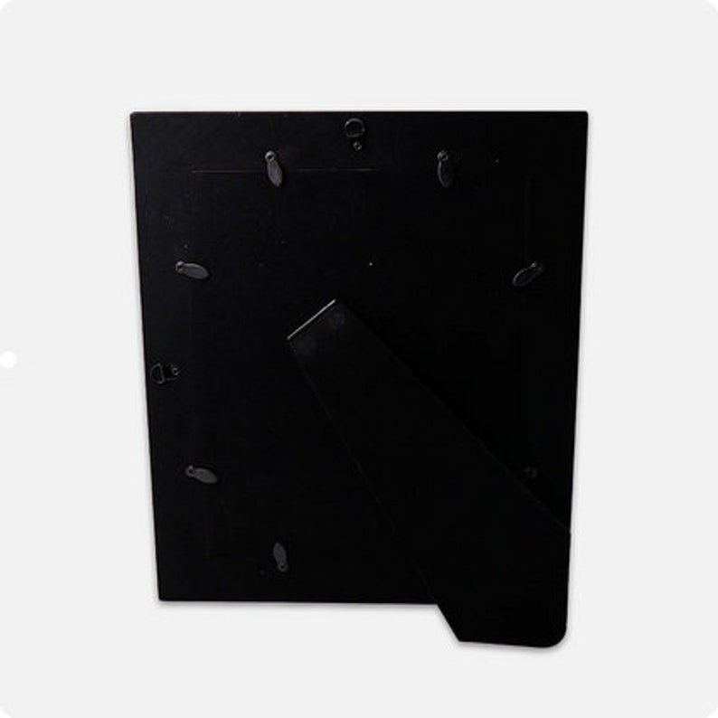 May include: A black picture frame back with a built-in easel stand. The back is made of a dark material and features several small, silver-colored clips to hold a picture in place. The easel is angled to allow the frame to stand upright.