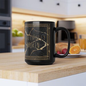 May include: Black ceramic mug with a gold marble design and the words "BEAUTIFULLY BROKEN" in gold lettering. The mug has a large handle and is sitting on a wooden surface.