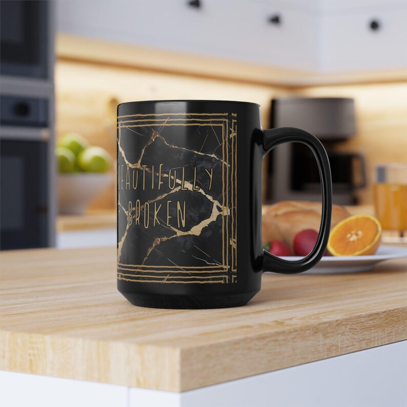 May include: Black ceramic mug with a gold marble design and the words "BEAUTIFULLY BROKEN" in gold lettering. The mug has a large handle and is sitting on a wooden surface.
